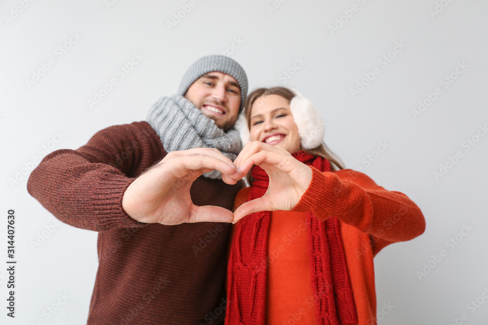 Happy couple in winter clothes making heart shape with their hands on light background