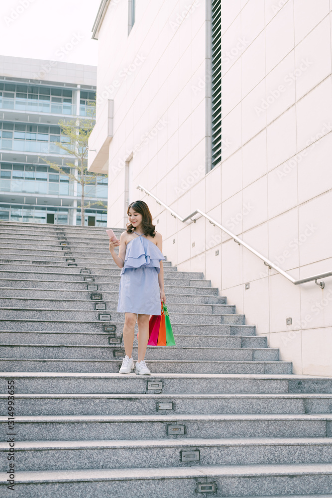 快乐的年轻亚洲女人用电话，带着五颜六色的商店包裹走下楼梯。