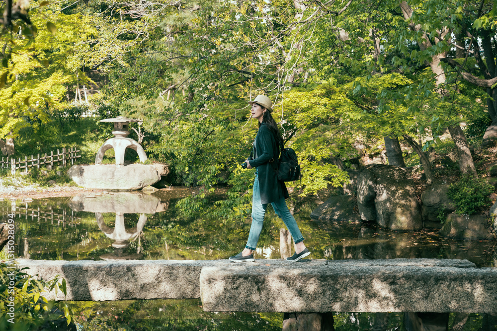 在绿色植物环绕的春天公园里行走的年轻女孩背包旅行者的全长。侧面