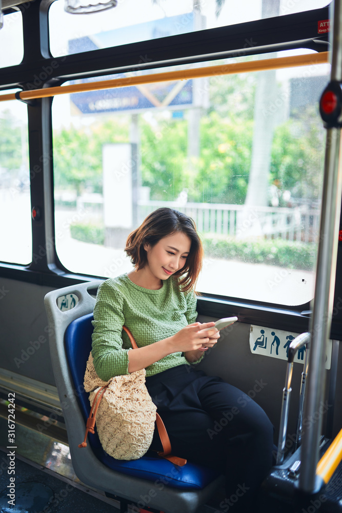 美丽快乐的年轻女子坐在城市公交车上，看着手机