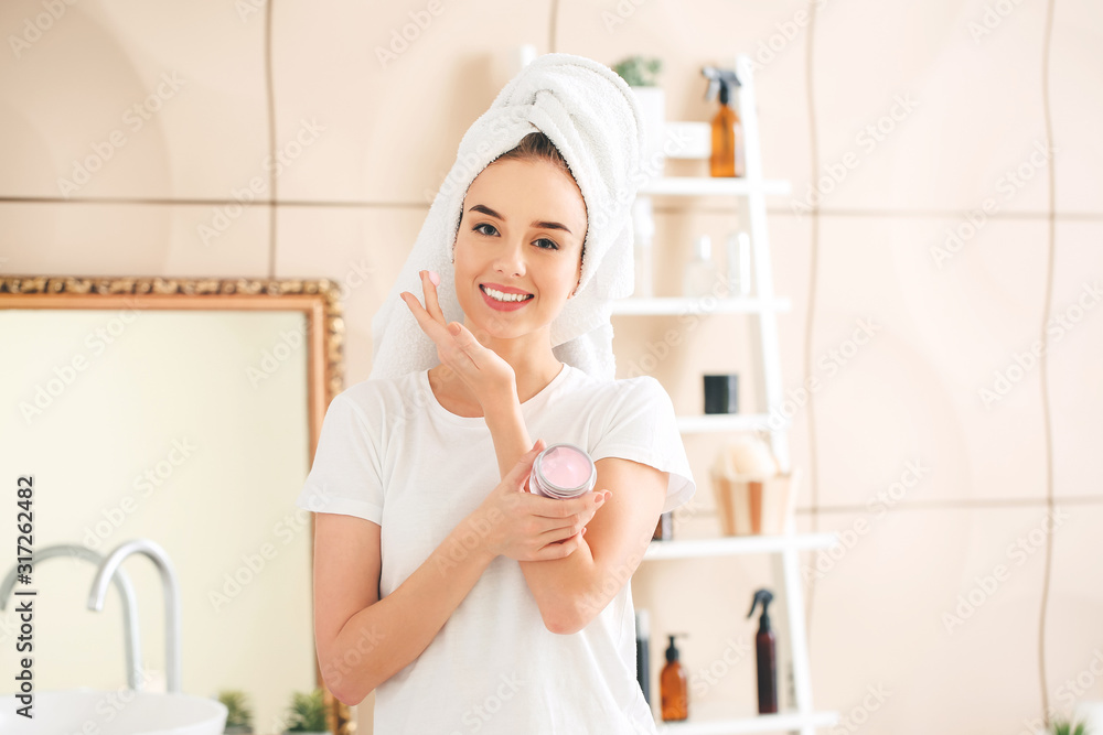 Beautiful young woman applying cream on her face in bathroom