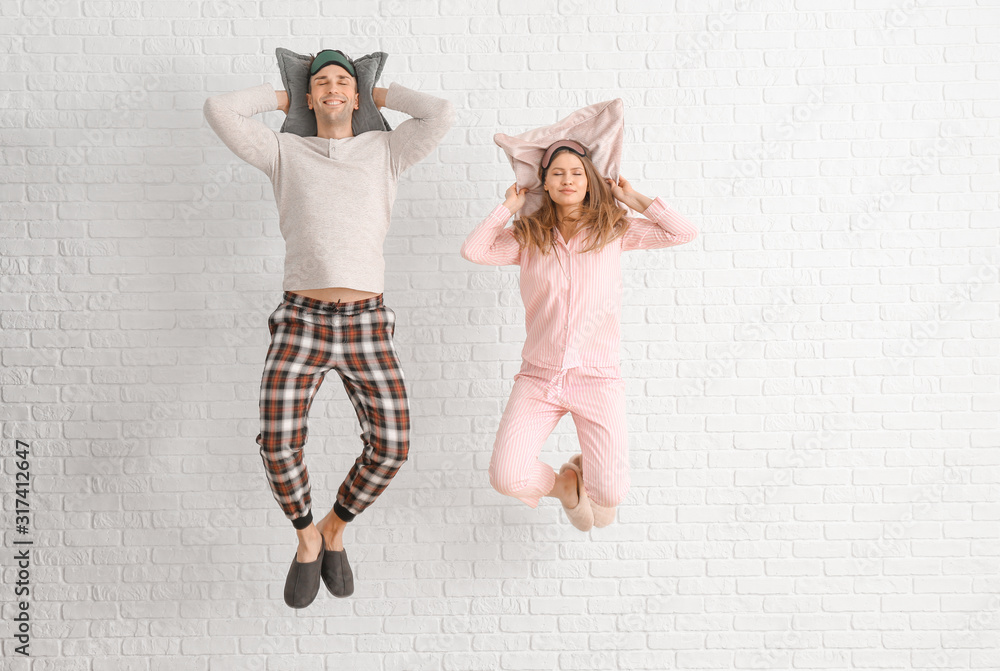 Jumping young couple in pajamas and with pillows on white brick background