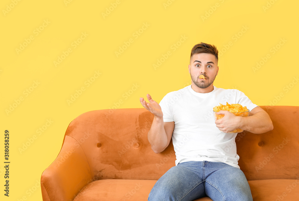 Shocked young man with tasty potato chips sitting on sofa near color wall