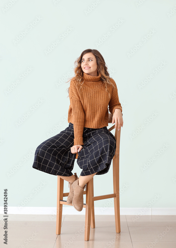 Beautiful young woman in warm sweater sitting on chair against light wall