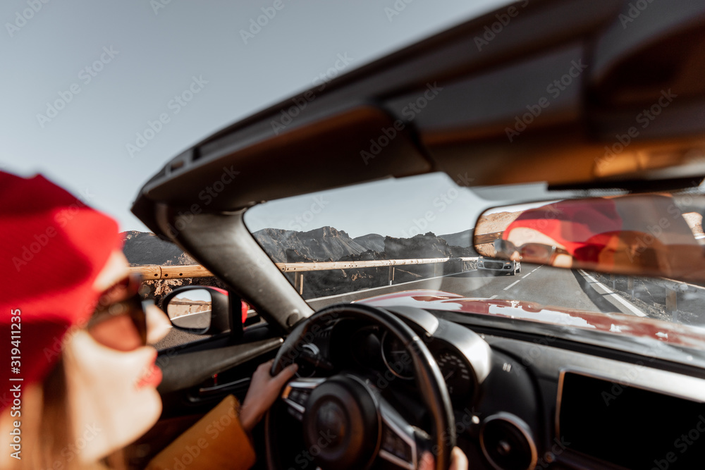 Happy woman in bright hat and jacket driving convertible car while traveling on the desert road on a