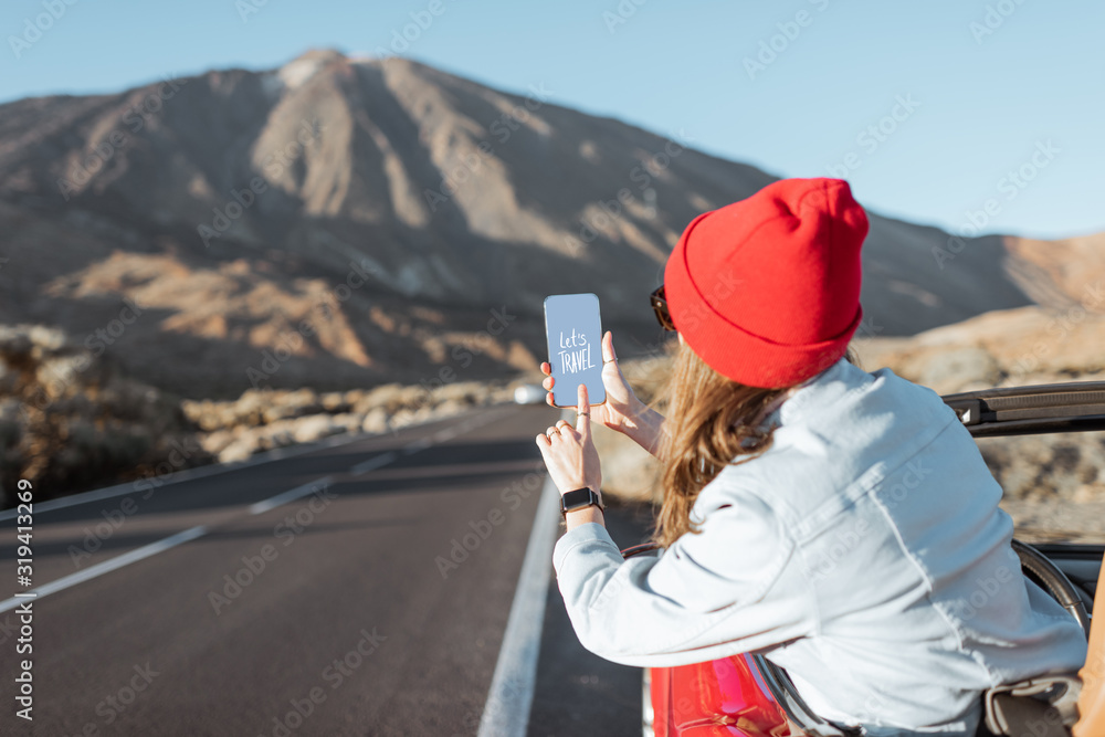 在火山谷上开车旅行的女人，从车窗探出头，手里拿着智能手机。