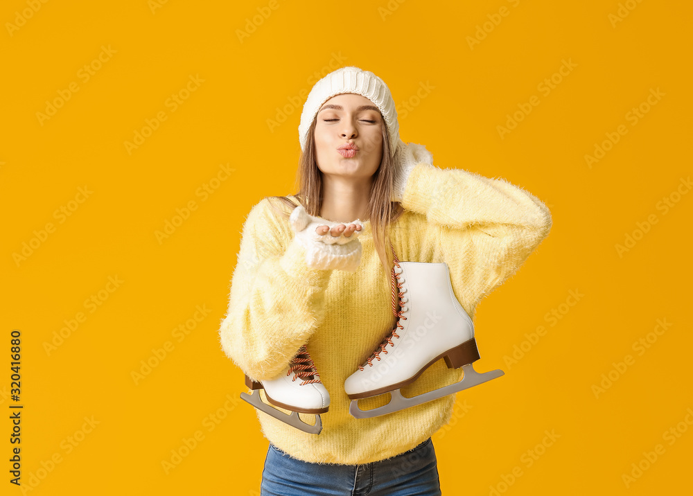 Beautiful young woman with ice skates blowing kiss on color background