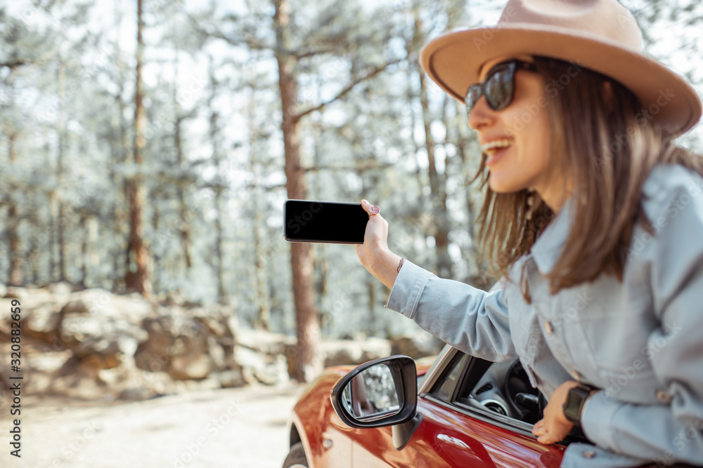 Woman traveling by car in the forest, leaning out of the car window and photographing with a smart p