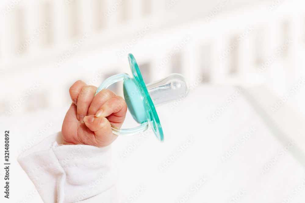 Little tiny infant boy hand lifted up in the air holding pacifier with baby crib on background