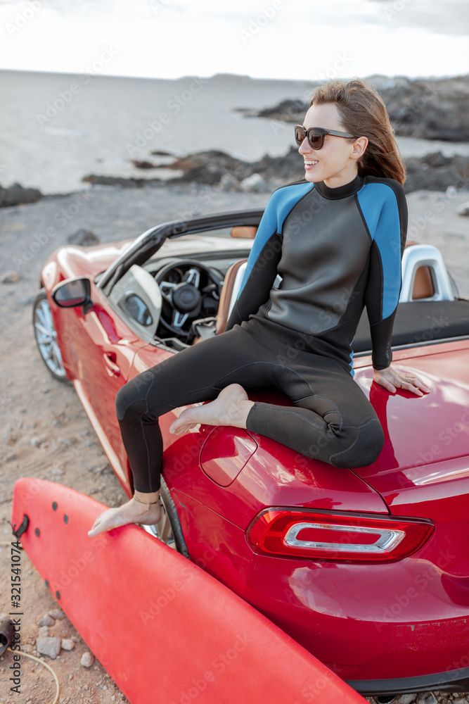 Portrait of a young woman surfer in swimsuit sitting with surfboard on the red cabriolet on the rock