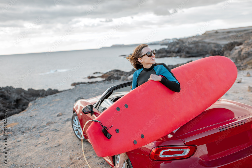 Portrait of a young woman surfer in swimsuit sitting with surfboard on the red cabriolet on the rock