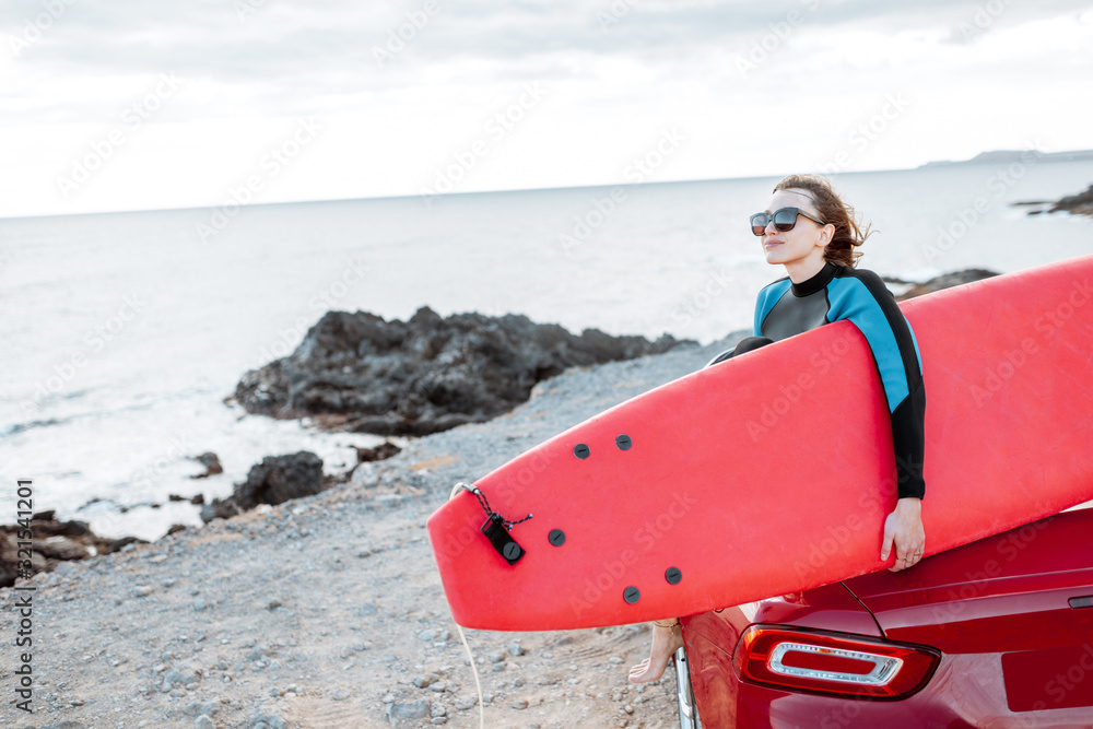 Portrait of a young woman surfer in swimsuit sitting with surfboard on the red cabriolet on the rock