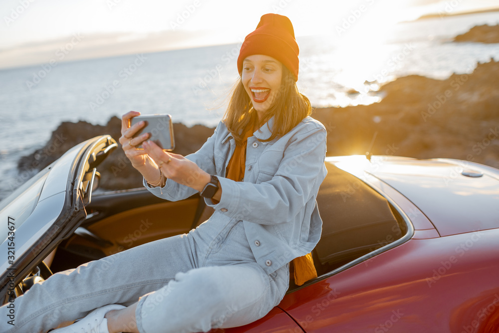 Young stylish woman photographing or vlogging on phone while traveling by car on the rocky coast nea