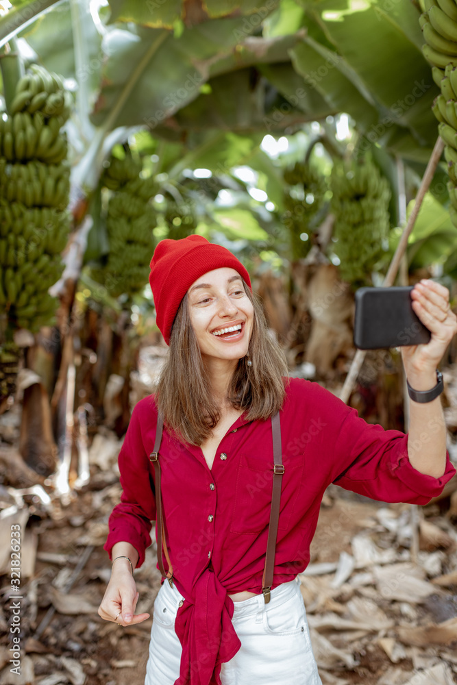 Woman as a tourist dressed in red exploring banana plantation, photographing or vlogging on phone. C