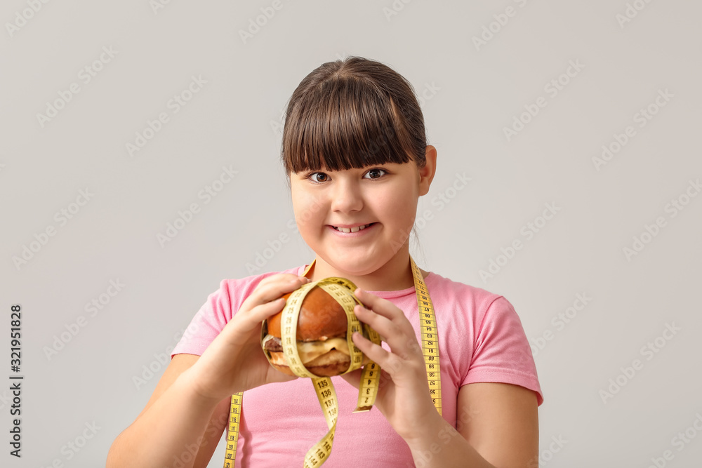 Overweight girl with unhealthy burger and measuring tape on light background