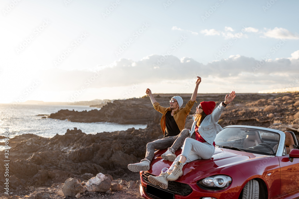 Young lovely couple enjoying landscapes, sitting together on a car hood, traveling by car on the roc