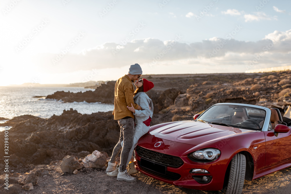 Young lovely couple kissing on the beach, traveling by car on the rocky ocean coast on a sunset. Car