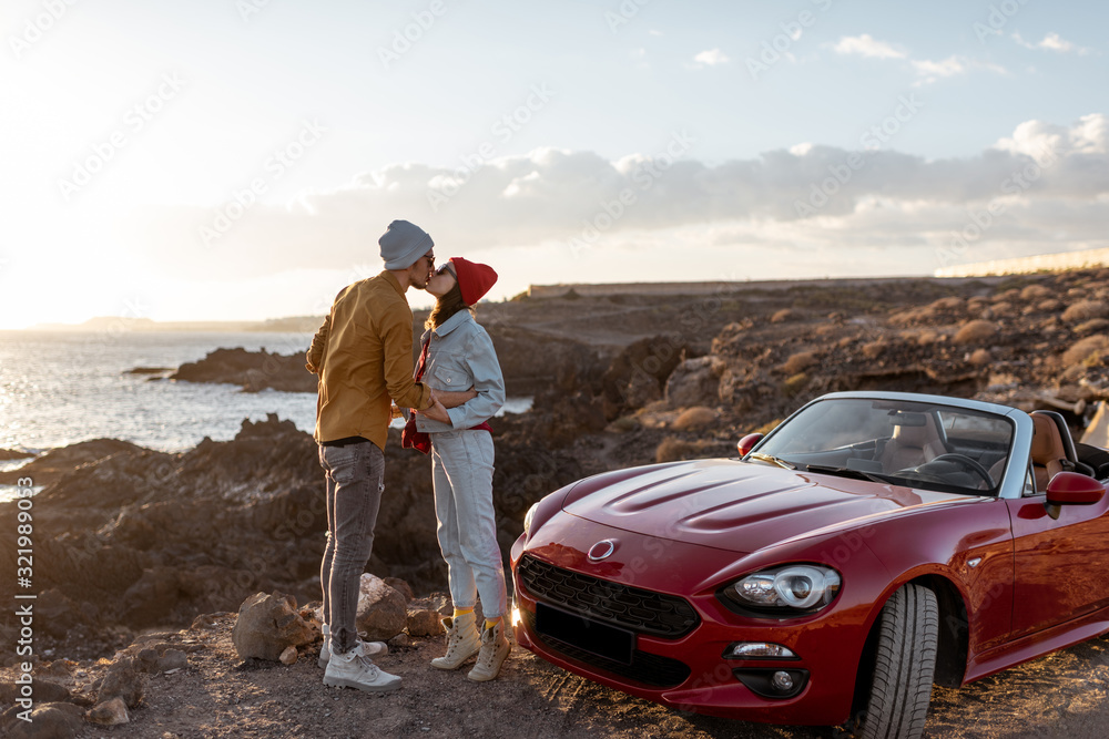 Young lovely couple kissing on the beach, traveling by car on the rocky ocean coast on a sunset. Car