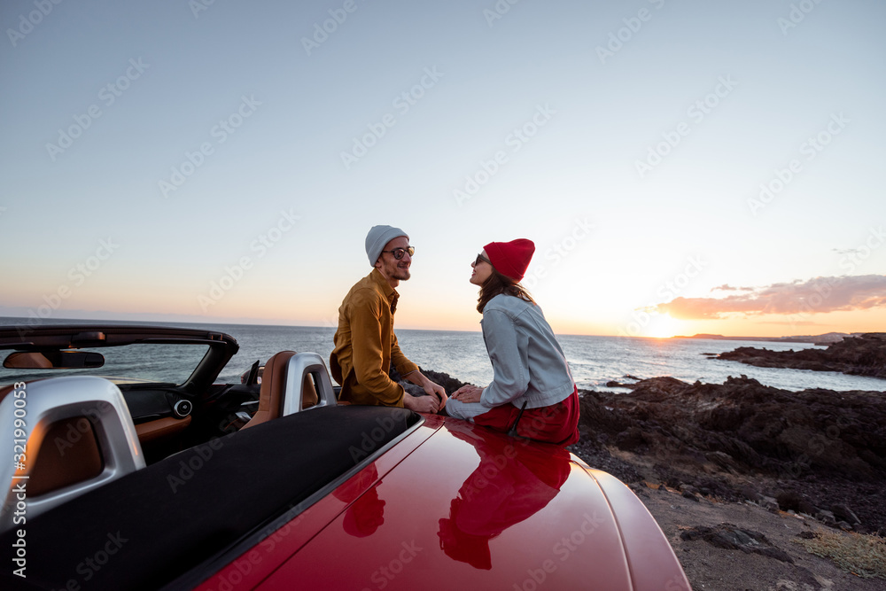 Portrait of a lovely couple dressed casually in hats sitting together on a car during a sunset, trav