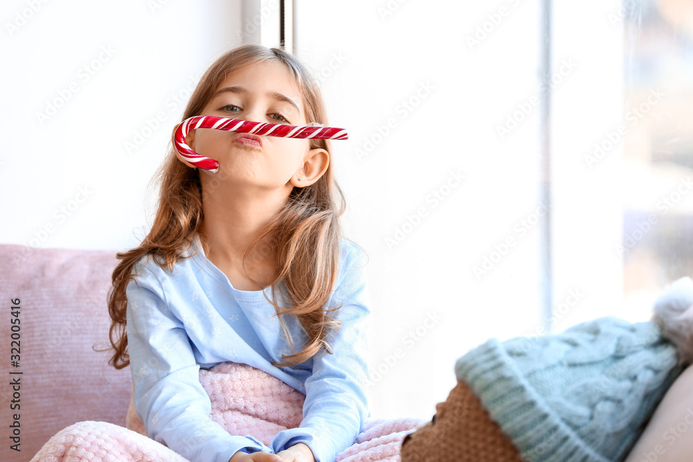 Cute little girl with Christmas candy cane on window sill at home