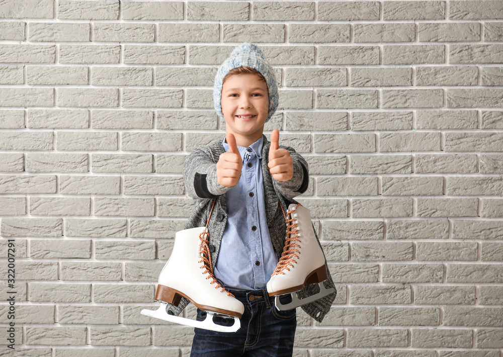 Cute little boy with ice skates showing thumb-up against brick background