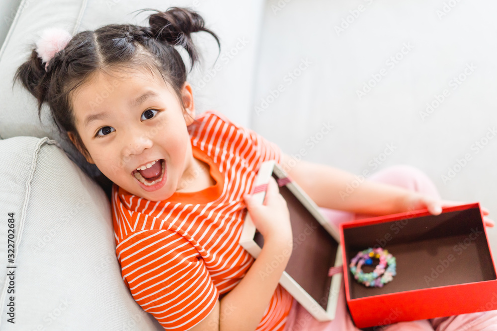 Little asian girl smile and excited and holding red gift box on sofa in living room background.child