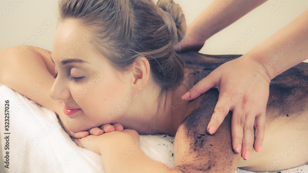 Relaxed young woman lying on spa bed for body scrubbing massage using traditional herbs and spices b