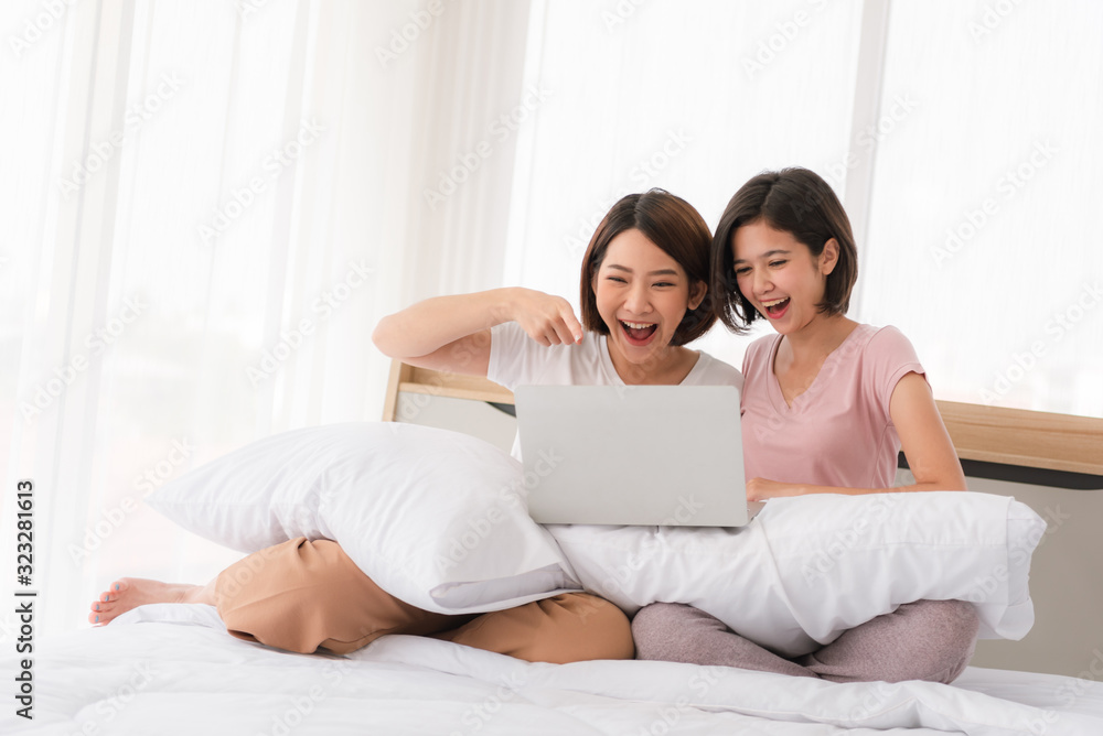 Happy casual two beautiful women working on a laptop sitting on the bed in the house.