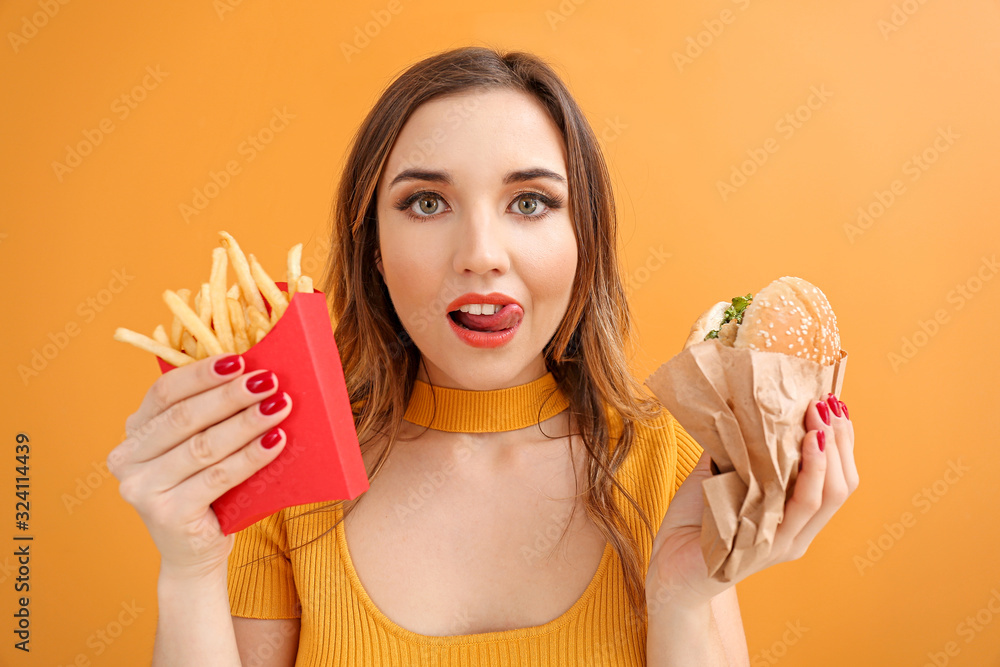 Portrait of beautiful young woman with burger and french fries on color background