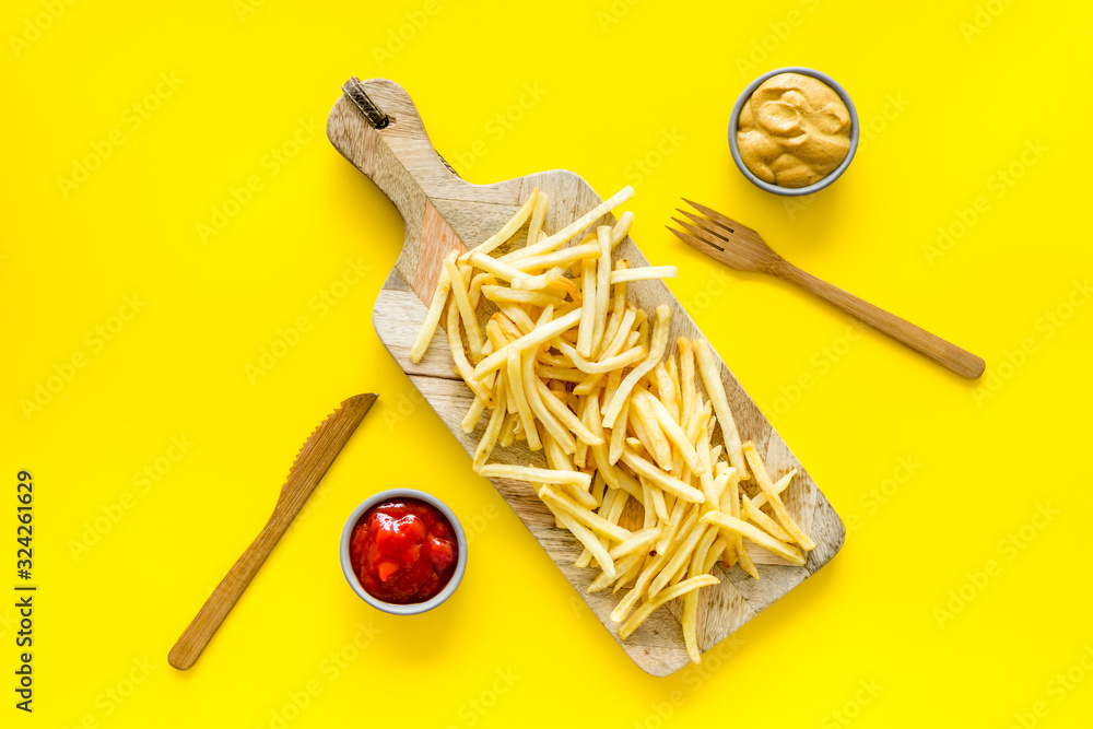 Fast food restaurant concept. French fries on cutting board near sauces on yellow table top-down