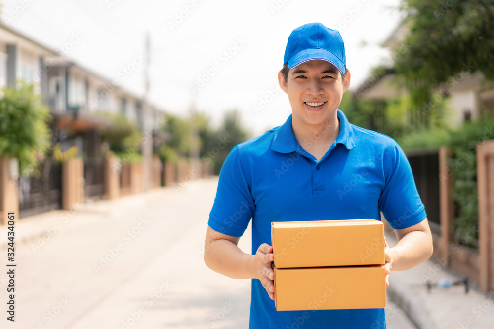 Asian delivery young man in blue uniform smile and holding pile of cardboard boxes in front house vi