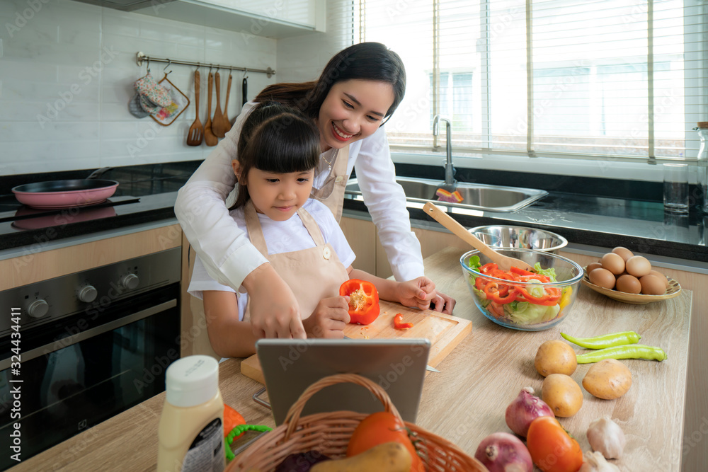 Portrait of beautiful Asian young woman and her daughter cooking salad for lunch using online intern