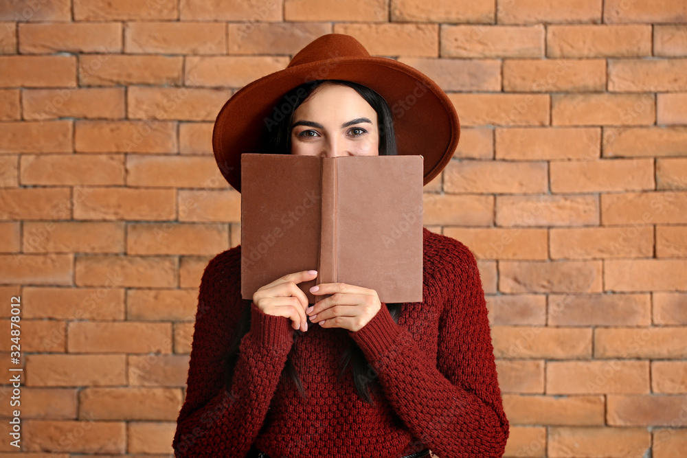 Beautiful young woman in warm sweater reading book on brick background