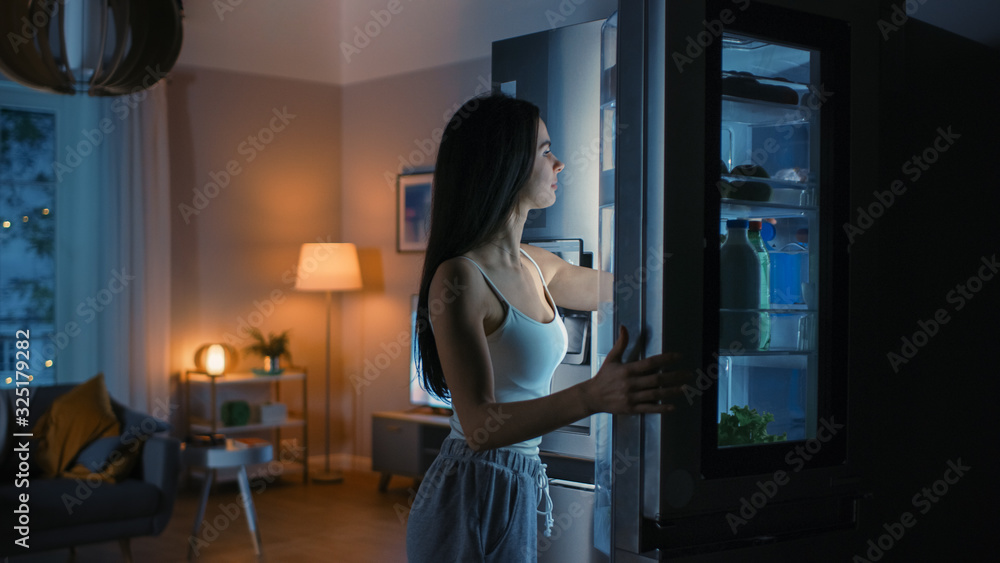 饥饿的美丽年轻女子晚上在厨房打开冰箱，拿出一块