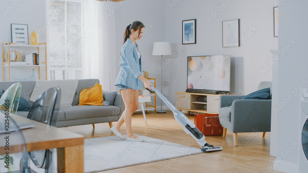 Young Beautiful Woman in Jeans Shirt and Shorts is Vacuum Cleaning a Carpet in a Bright Cozy Room at