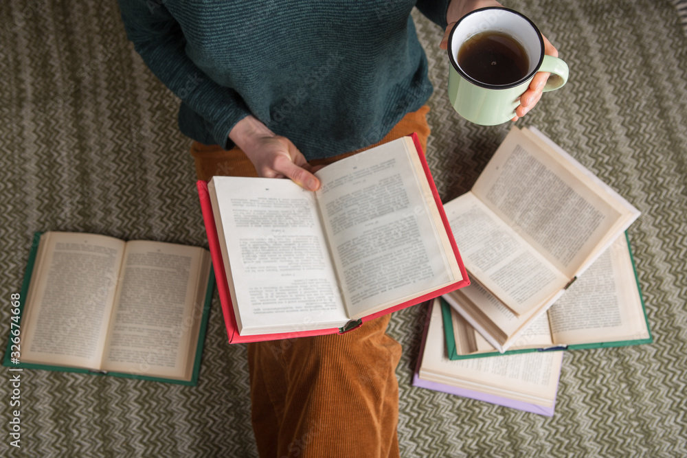 Young woman is sitting on a carpet and reading a book, holding a cup of tea