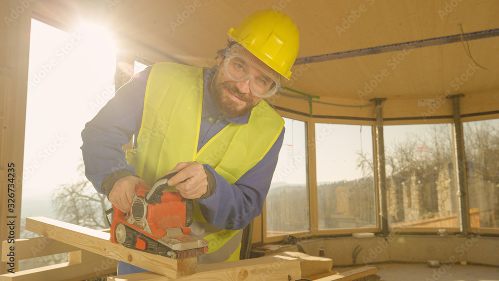 PORTRAIT, LENS FLARE: Construction worker smiles while sanding a wooden beam.