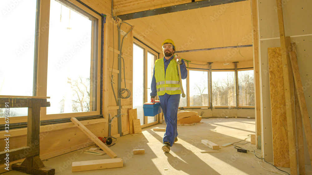 CLOSE UP: Contractor in blue overalls carries a toolbox and ladder across room