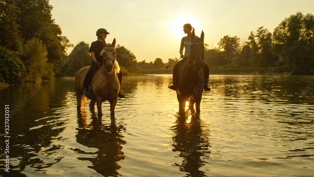 CLOSE UP: Two horses standing in shallow river with riders at golden sunrise