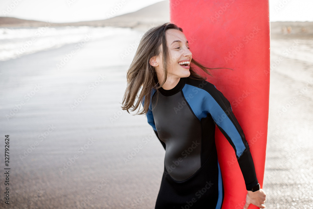 Portrait of a young joyful woman in wetsuit standing with red surfboard on the ocean beach during a 