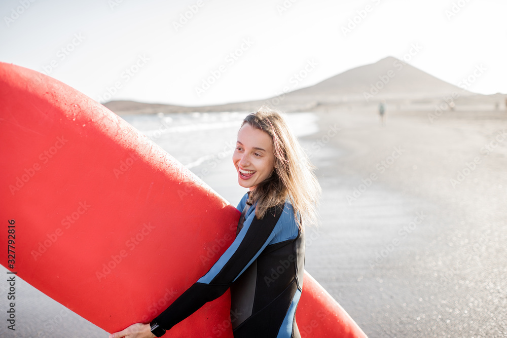 一位穿着潜水服、拿着红色冲浪板站在海滩上的年轻快乐女性的肖像