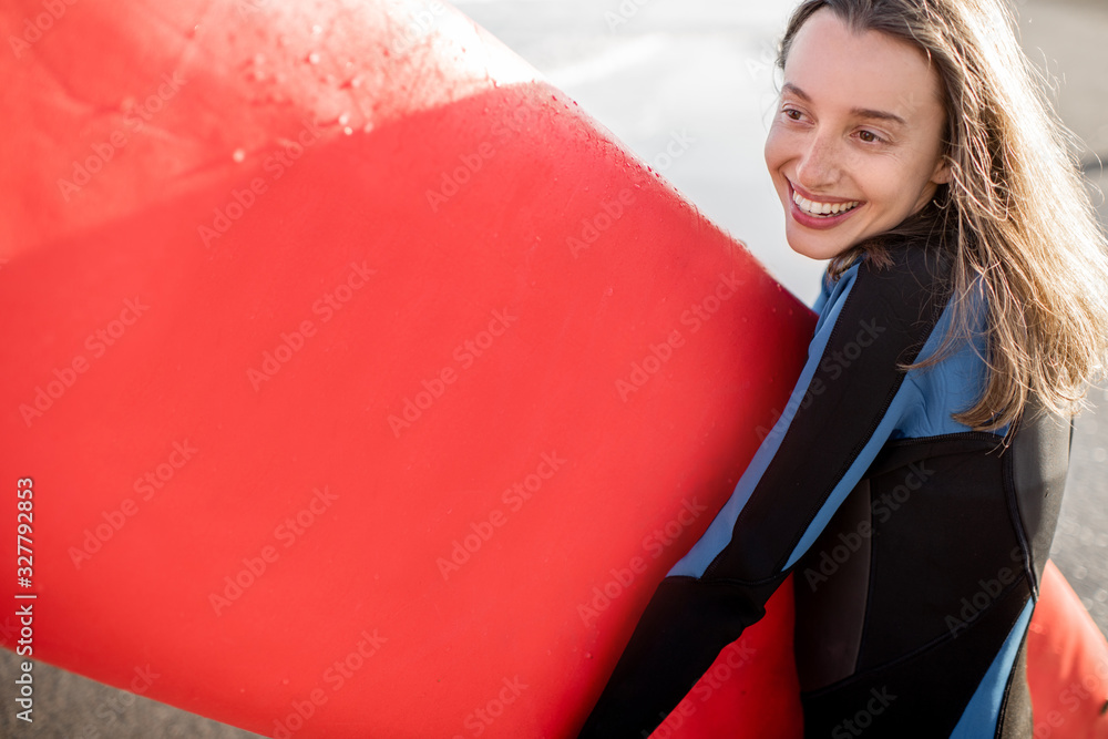 Portrait of a young joyful woman in wetsuit standing with red surfboard on the ocean beach during a 