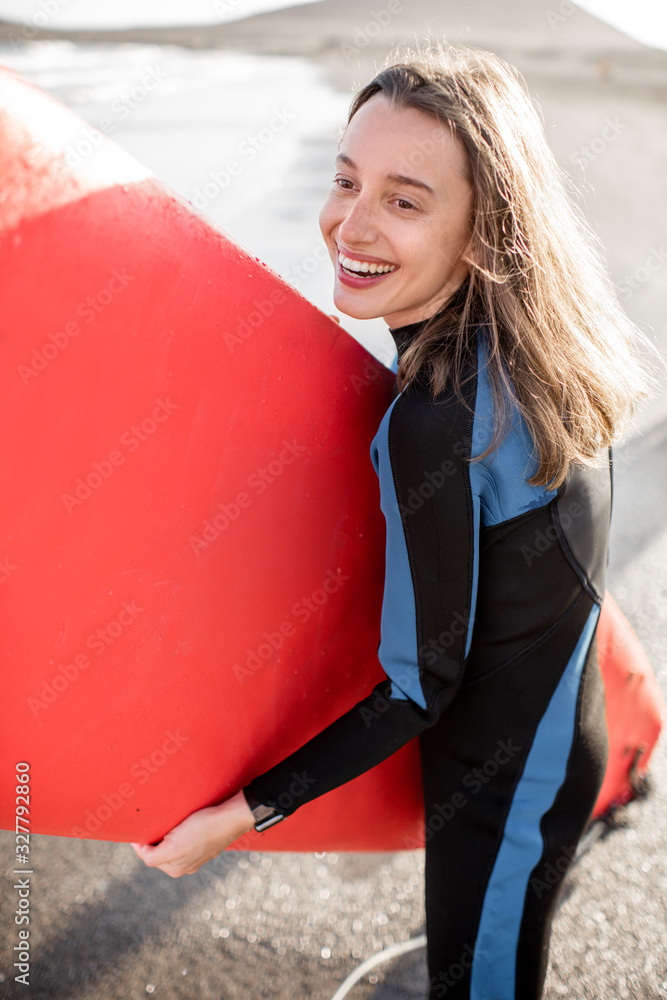 一位穿着潜水服、拿着红色冲浪板站在海滩上的年轻快乐女性的肖像