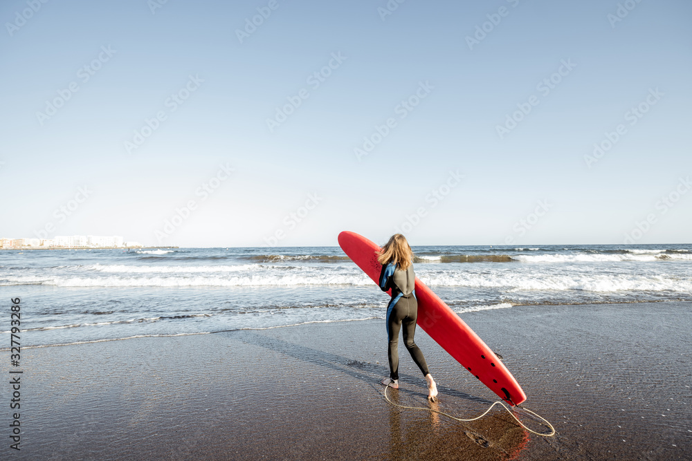 Young surfer in wetsuit walking with red surfboard on the beautiful ocean beach during a sunset, wid