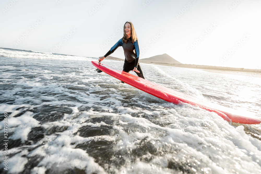 Young surfer in wetsuit getting on surfboard, catching water flow near the beach during a sunset. Wa