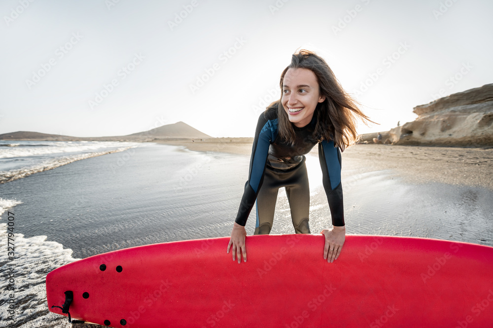 Portrait of a young joyful woman in wetsuit standing with red surfboard on the ocean beach during a 