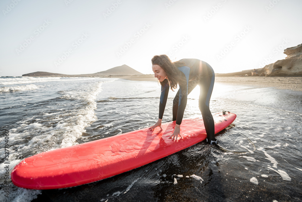 Young surfer in wetsuit getting on surfboard, catching water flow near the beach during a sunset. Wa