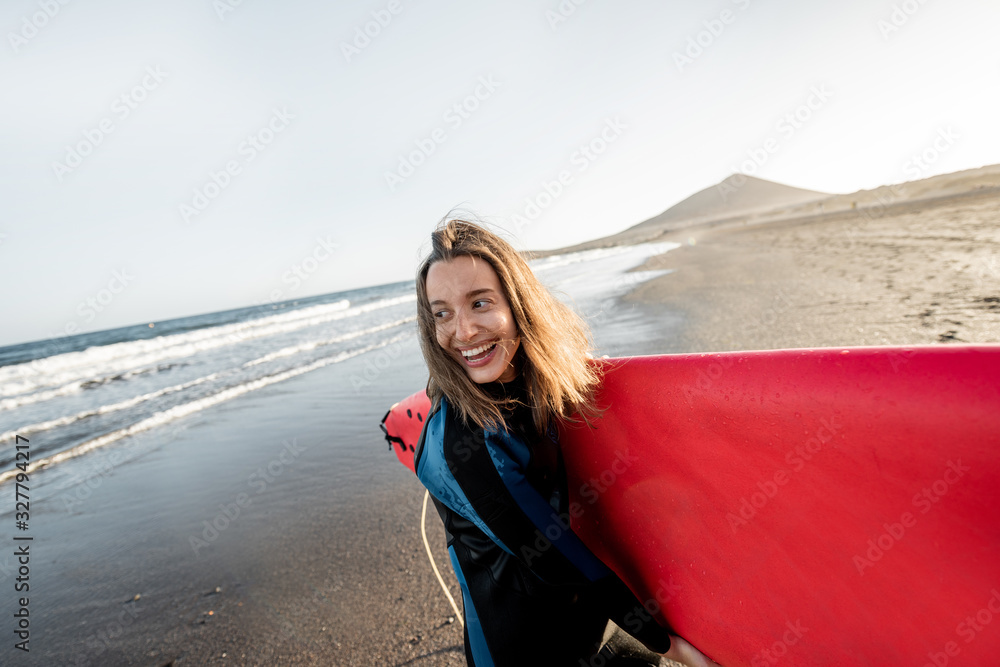 Portrait of a young joyful woman in wetsuit carrying surfboard on the ocean beach during a sunset. W