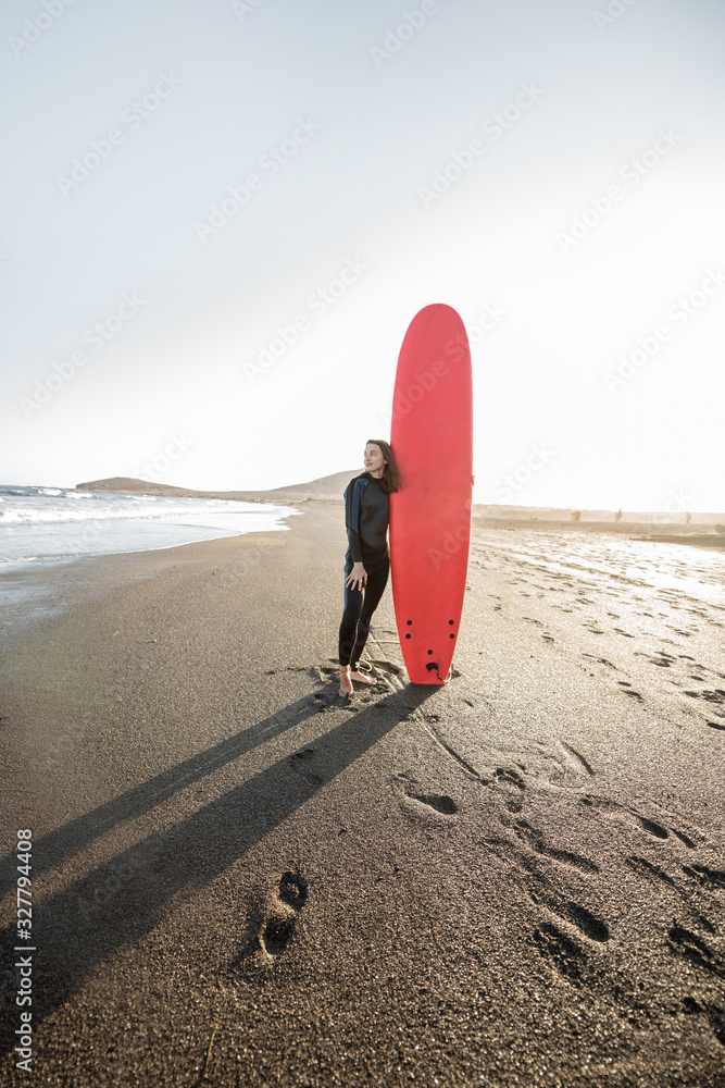 Portrait of a young woman in wetsuit standing with red surfboard on the ocean beach during a sunset.