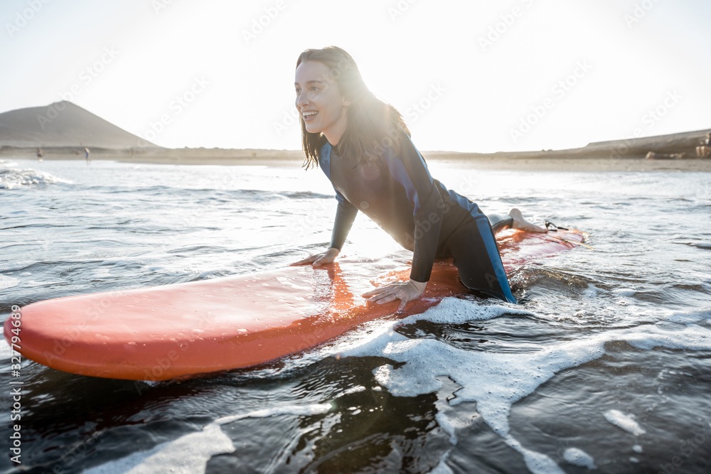 Young surfer in wetsuit getting on surfboard, catching water flow near the beach during a sunset. Wa